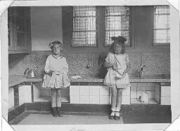 Twee meisjes van een Montessorischool staan voor het keukentje in een klaslokaal. 191?