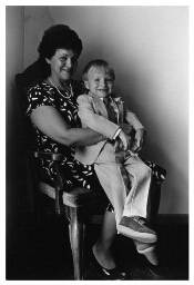 Een vrouw met op haar schoot een jongen, die in pak is gestoken voor een trouwerij. 1985