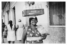 Een vrouw draagt een krat met cola flesjes op haar hoofd. 1984