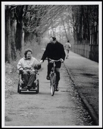 Man op een fiets en gehandicapte vrouw in een rolstoel rijden hand in hand over het fietspad 1995