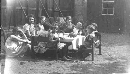 Groep kinderen zit buiten aan tafel te eten. 1926