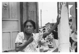 Een oudere vrouw staat tegen een pilaar, achter haar zie je de drukte van het dorpsleven. 1984