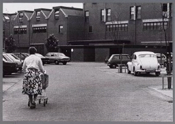 Vrouw loopt met winkelkarretje over parkeerterrein 1982