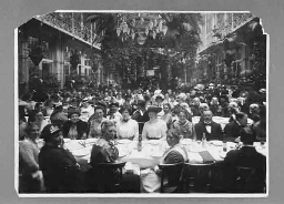 Grote groep vrouwen en mannen aan lange eettafels t.g.v 1916