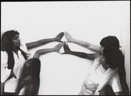 Vier vrouwen met verschillende huidskleuren vormen met hun handen twee keer het Yoni-teken (driehoek-teken) op een witte muur