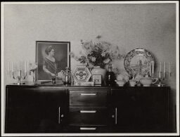 Interieur met dressoir waarop cadeaus  ter ere van het 25-jarig jubileum van de Vereniging van Huisvrouwen staan uitgestald. 1937