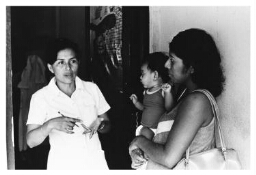 Vrouw en haar kind wachten op de dokter, de verpleegster komt even kijken. 1984