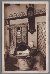 Vrouw in klederdracht zit bij een wieg in kamer. 1913