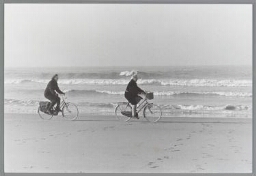 Twee vrouwen fietsen langs de branding van de zee. 1990