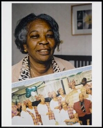 Portret van voormalig verpleegkundige Elisabeth Bottse met een foto van de seniorendansgroep Amsterdam Zuidoost 2002
